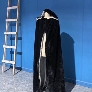 May include: A floor-length black velvet cloak featuring a hood and a contrasting light gray lining. The garment is displayed on a mannequin and includes a tie closure at the neck, showcasing a soft, textured fabric with a slight sheen throughout.