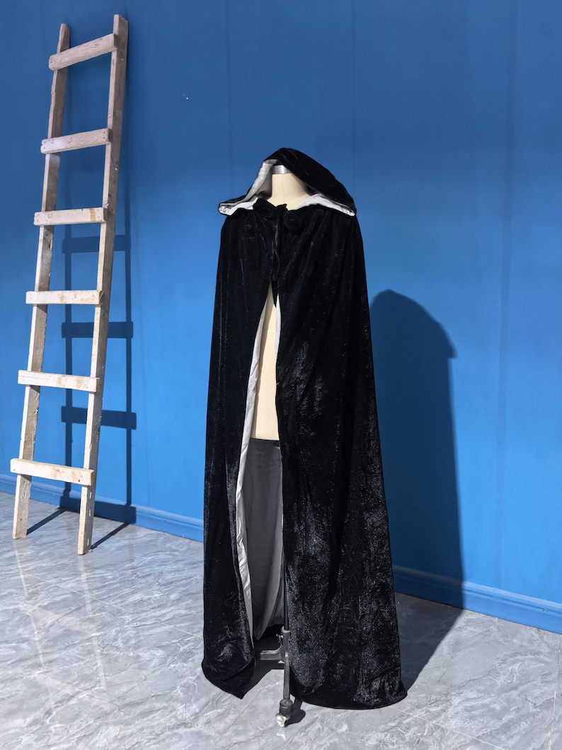 May include: A floor-length black velvet cloak featuring a hood and a contrasting light gray lining. The garment is displayed on a mannequin and includes a tie closure at the neck, showcasing a soft, textured fabric with a slight sheen throughout.