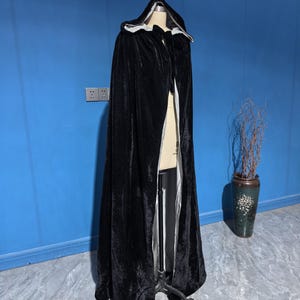 May include: A floor-length black velvet hooded cloak featuring a light gray interior lining. The garment is draped over a mannequin, showcasing a deep hood, a full-length silhouette, and a smooth, dark exterior texture with a contrasting lighter lining.