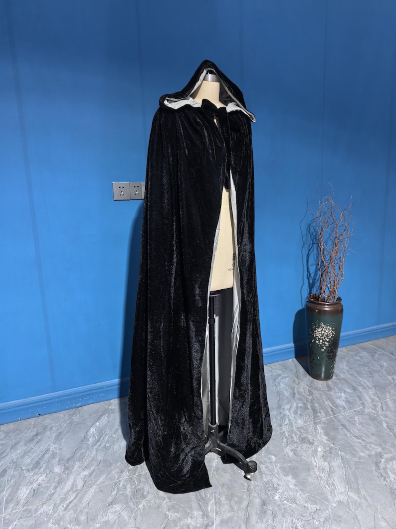 May include: A floor-length black velvet hooded cloak featuring a light gray interior lining. The garment is draped over a mannequin, showcasing a deep hood, a full-length silhouette, and a smooth, dark exterior texture with a contrasting lighter lining.