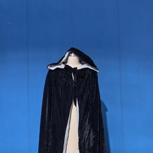 May include: A black velvet hooded cloak featuring a white lining. The garment includes a hood and a tie closure at the neck, draping down from the shoulders. The exterior fabric has a soft, reflective texture, while the interior is a solid white.