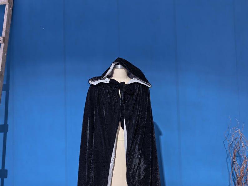 May include: A black velvet hooded cloak featuring a white lining. The garment includes a hood and a tie closure at the neck, draping down from the shoulders. The exterior fabric has a soft, reflective texture, while the interior is a solid white.