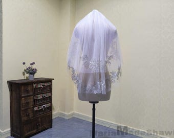 May include: A sheer white bridal veil with silver floral embroidery is displayed on a mannequin. The veil has a delicate, flowing appearance. The mannequin is on a black stand. The background is a pale yellow wall.