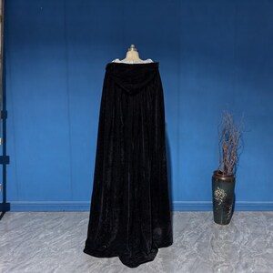 May include: A floor-length black velvet hooded cloak displayed on a mannequin. The garment features a deep hood and a full, flowing silhouette that extends to the floor, crafted from a dark, soft-textured fabric with a subtle sheen.