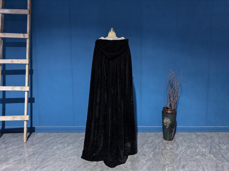 May include: A floor-length black velvet hooded cloak displayed on a mannequin. The garment features a deep hood and a full, flowing silhouette that extends to the floor, crafted from a dark, soft-textured fabric with a subtle sheen.