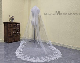 May include: A long, sheer white bridal veil with a delicate, ornate lace trim along the bottom edge. The veil is displayed on a mannequin, showcasing its length and design. The text "MariaMadeShawl" is visible in the background.