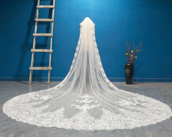 Puede incluir: Un velo de novia blanco largo con intrincados detalles de encaje en los bordes y la cola. El velo está drapeado sobre un maniquí, mostrando su longitud y diseño. El fondo es una pared azul sólida.