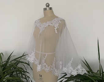 May include: White sheer lace bridal cape with a scalloped edge and floral lace detailing. The cape is on a white dress form.