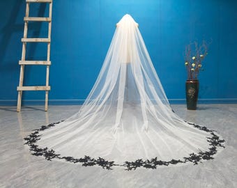 May include: A long, white bridal veil with a black floral lace trim along the bottom edge. The veil is displayed on a mannequin against a blue background. A wooden ladder and a decorative vase are also visible.