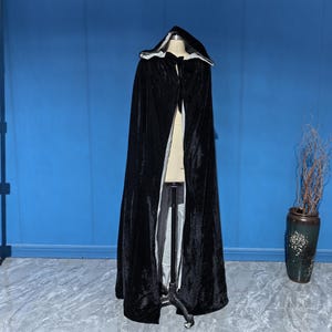 May include: A floor-length black velvet cloak featuring a deep hood and a contrasting light-colored interior lining. The garment is secured at the neck with a simple tie closure and is displayed on a mannequin stand, showcasing its full length.