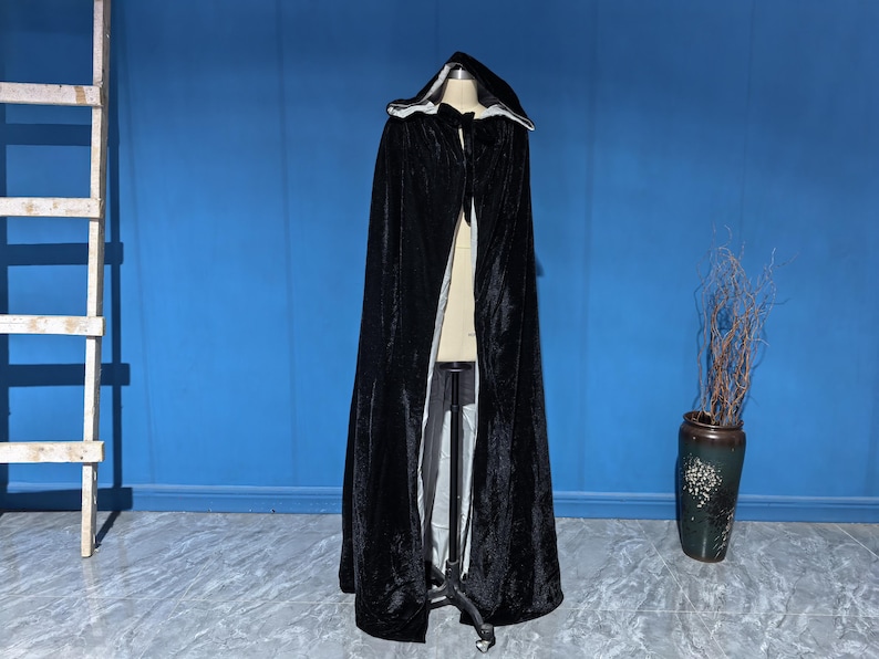 May include: A floor-length black velvet cloak featuring a deep hood and a contrasting light-colored interior lining. The garment is secured at the neck with a simple tie closure and is displayed on a mannequin stand, showcasing its full length.