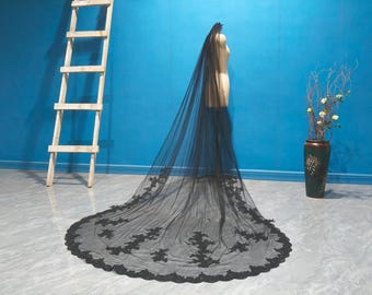 May include: A black bridal veil with intricate floral lace detailing along the edges. The veil is displayed on a mannequin against a blue backdrop. A wooden ladder and a decorative vase are also visible.
