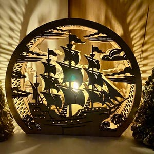 Pode incluir: Lâmpada de madeira, circular, com tema náutico, com um recorte detalhado de um veleiro com velas esvoaçantes, contra um fundo de nuvens. A lâmpada é iluminada por dentro, projetando sombras intrincadas. Dois objetos decorativos em forma de árvore ladeiam a lâmpada.