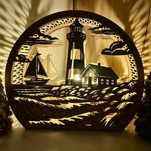Puede incluir: Una escena de faro circular de madera con un velero, una casa y un faro. El diseño está recortado, permitiendo que la luz brille. El faro es blanco y negro, y la casa tiene una pequeña luz en su interior. La escena está ambientada en una playa con olas.