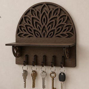 May include: A dark brown wooden wall-mounted key holder featuring a semi-circular top with an intricate laser-cut floral pattern. The unit includes a small horizontal shelf supported by two decorative brackets and five hooks, each holding a metal key.