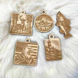 Pode incluir: Cinco ornamentos de madeira com designs cortados a laser. Os ornamentos apresentam um veado na floresta, uma cena de montanha, um peixe, uma águia com uma bandeira americana e um farol com ondas.