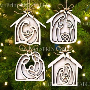 May include: Four wooden Christmas ornaments in the shape of a house, each featuring a layered Nativity scene. The ornaments are light-colored with a natural wood grain, and are suspended by twine. The ornaments are hanging on a green Christmas tree.