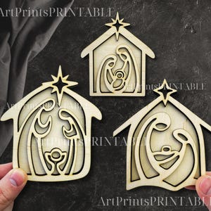 May include: Three wooden nativity scene ornaments. Each ornament features a house shape with a star on top and a cutout design depicting Mary, Joseph, and baby Jesus. The ornaments are made of light-colored wood.
