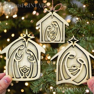 May include: Three wooden Christmas ornaments in the shape of a house, each featuring a nativity scene cutout. The ornaments are light brown with a star at the top and are hanging on a Christmas tree.