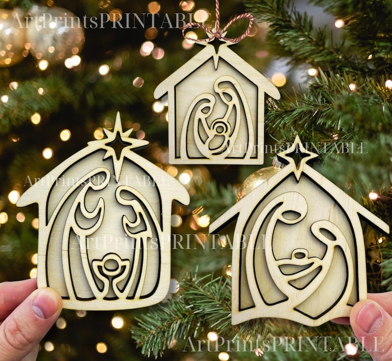 May include: Three wooden Christmas ornaments in the shape of a house, each featuring a nativity scene cutout. The ornaments are light brown with a star at the top and are hanging on a Christmas tree.