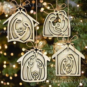 May include: Four wooden Christmas ornaments depicting the Nativity scene, hanging from twine. The ornaments are in the shape of a house or a rounded shape, with a star at the top. The ornaments are made of light-colored wood.