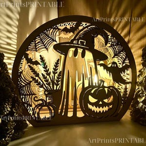 May include: Wooden Halloween-themed decorative light with a circular frame. The design features a ghost wearing a witch hat, a jack-o'-lantern, bats, and spiderwebs. The text "ArtPrintsPRINTABLE" is visible.