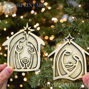 May include: Two wooden Christmas ornaments in the shape of a nativity scene. Each ornament features a star on top of a house-shaped frame, with the holy family inside. The ornaments are held in front of a Christmas tree.