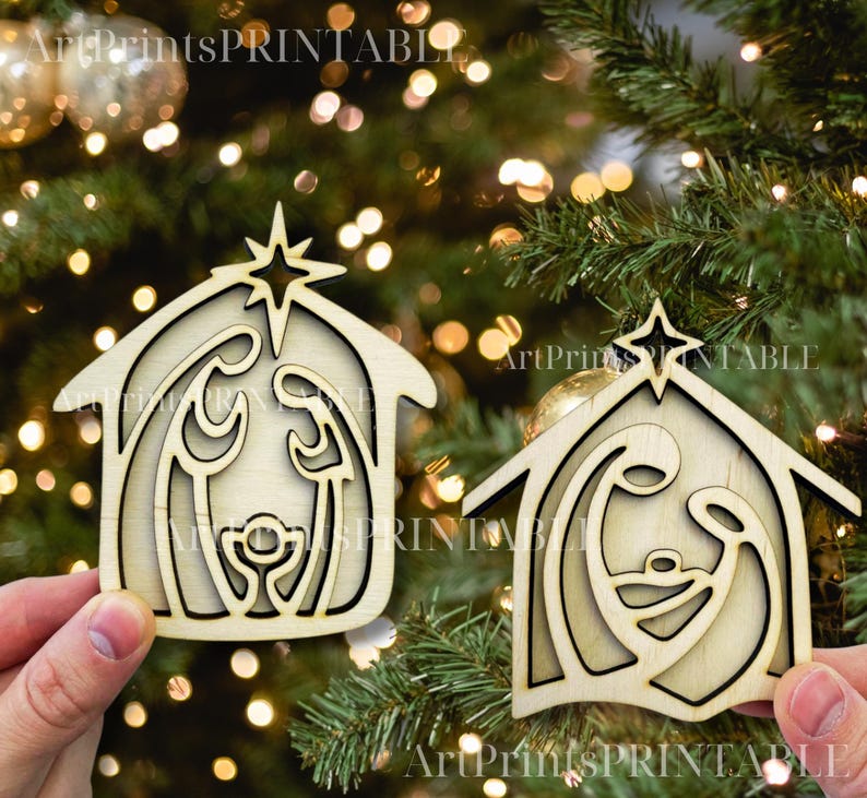 May include: Two wooden Christmas ornaments in the shape of a nativity scene. Each ornament features a star on top of a house-shaped frame, with the holy family inside. The ornaments are held in front of a Christmas tree.
