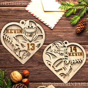 Puede incluir: Dos adornos de madera en forma de corazón con un diseño de jugador de fútbol americano. Los adornos están cortados con láser y presentan los nombres "Kevin" y los números "13" y "14".