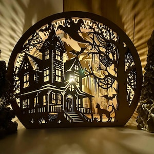 Puede incluir: Una luz decorativa de madera con temática de Halloween. El diseño presenta una casa embrujada, murciélagos, un gato y árboles, todos intrincadamente recortados. La luz brilla a través de los recortes, creando un ambiente espeluznante. El color general es marrón.