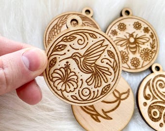 May include: Five wooden ornaments with laser-cut designs. The ornaments feature a hummingbird, a bee, flowers, and swirls. The ornaments are all round and have a natural wood finish.