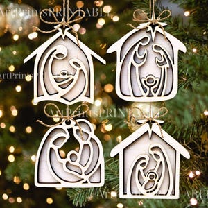 May include: Four wooden Christmas ornaments in the shape of a house, each featuring a nativity scene cutout. The ornaments are light-colored with a natural wood grain, and are suspended by twine. The background is a blurred Christmas tree.