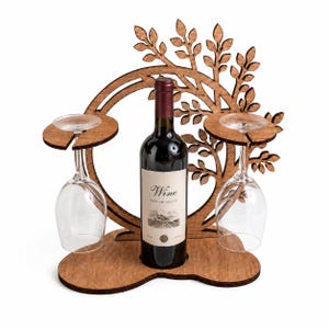 Puede incluir: Un soporte de madera marrón para botella de vino y copas con un marco circular y diseño de ramas de árbol. El soporte incluye una ranura central para una botella de vino y dos plataformas laterales con ranuras para colgar dos copas.