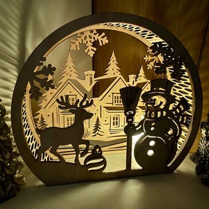 May include: A wooden Christmas scene with a snowman holding a broom, a deer, a house, and snowflakes. The scene is illuminated from behind, creating a warm glow.