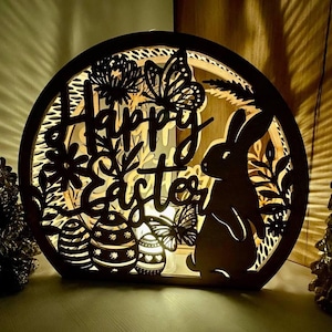 Op de afbeelding: Een houten paasdecoratie met een konijn, eieren, vlinders en de woorden "Happy Easter" uitgesneden. De decoratie wordt van achteren verlicht, waardoor een warme gloed ontstaat.