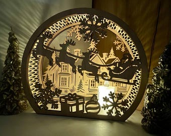 May include: A wooden Christmas light-up decoration featuring a silhouette of Santa Claus in a sleigh pulled by reindeer, flying over a house with snow-covered trees and snowflakes. The decoration is illuminated by a warm white light.
