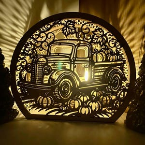 May include: A wooden, circular fall-themed decoration featuring a vintage pickup truck carrying pumpkins. The design is intricately cut out, with pumpkins and foliage surrounding the truck. The piece is illuminated from within, creating a warm glow.