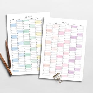 May include: Two 2026 yearly planners with a colorful grid layout, one in pastel colors and the other in cool tones. Two pencils and a binder clip are on the white surface. The planners are for scheduling and organization.