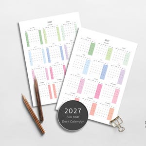 May include: Two white sheets of paper displaying a 2027 full-year calendar with months organized in rows and color-coded columns. Two wooden pencils and a silver metal binder clip are placed near a circular black label reading 2027 Full Year Desk Calendar.