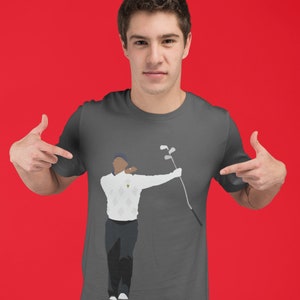 May include: A charcoal gray t-shirt featuring a graphic of a golfer in a white sweater and black pants, holding a golf club. The golfer is depicted in a mid-swing pose. The background is a solid red.