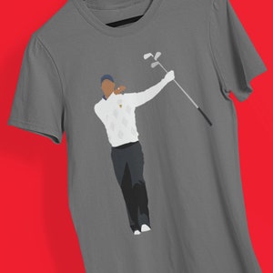 May include: Gray t-shirt featuring a graphic of a golfer in a white sweater with a diamond pattern, dark pants, and a blue cap. The golfer is mid-swing, holding a golf club. The shirt is hanging on a wooden hanger against a red background.