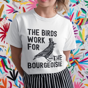 Puede incluir: Camiseta blanca con un gráfico en blanco y negro de un cuervo con un sombrero de copa. El texto en la camiseta dice "The Birds Work For The Bourgeoisie".