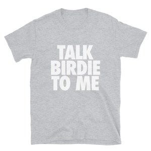 May include: A light gray t-shirt with the phrase "TALK BIRDIE TO ME" printed in large, white, block letters. The shirt is made of a soft material and has short sleeves and a crew neck. The text is centered on the front of the shirt.