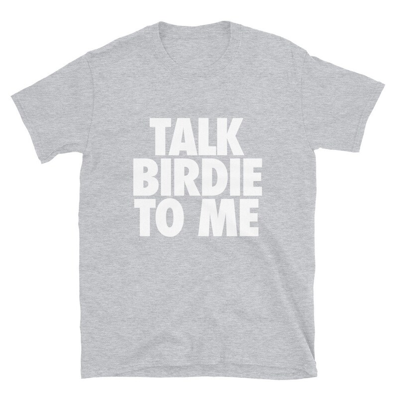 May include: A light gray t-shirt with the phrase "TALK BIRDIE TO ME" printed in large, white, block letters. The shirt is made of a soft material and has short sleeves and a crew neck. The text is centered on the front of the shirt.
