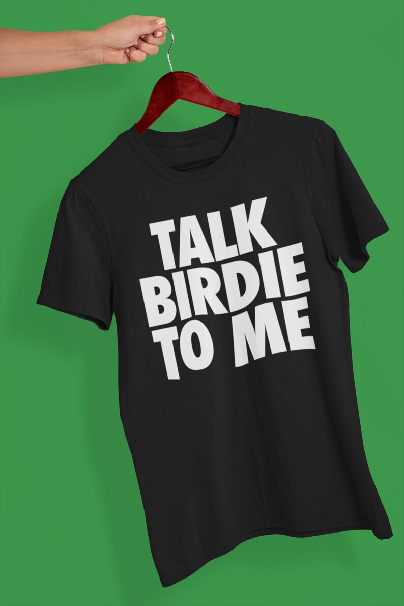 May include: Black t-shirt with the phrase "TALK BIRDIE TO ME" in bold white letters. The shirt is hanging on a wooden hanger against a green background. The text is centered and takes up most of the shirt's front.