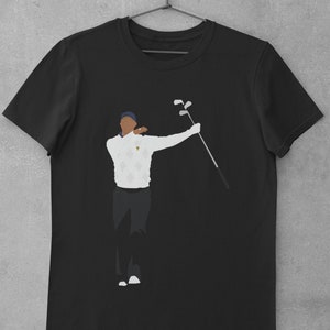 May include: Black t-shirt featuring a stylized illustration of a golfer in a white sweater with a diamond pattern, dark pants, and a blue cap. The golfer is holding golf clubs. The shirt is hanging on a hanger.