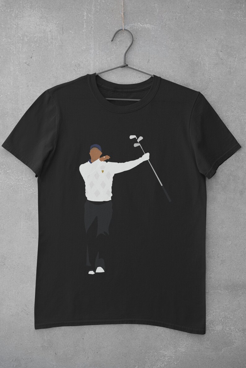 May include: Black t-shirt featuring a stylized illustration of a golfer in a white sweater with a diamond pattern, dark pants, and a blue cap. The golfer is holding golf clubs. The shirt is hanging on a hanger.