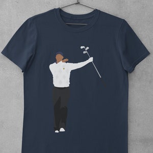 May include: Navy blue t-shirt featuring a stylized illustration of a golfer in a white sweater with a diamond pattern, black pants, and a blue cap. The golfer is holding golf clubs. The shirt is hanging on a metal hanger.