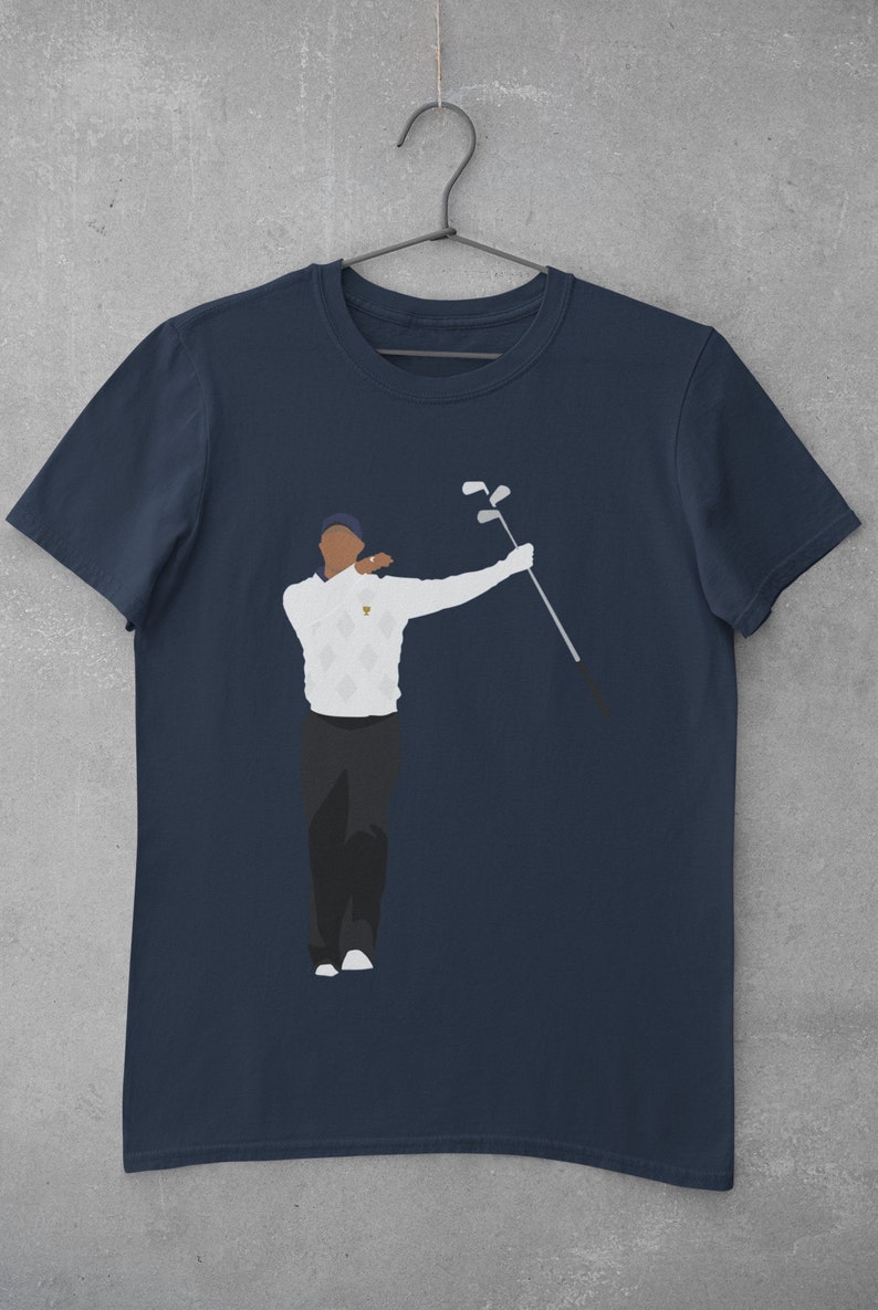 May include: Navy blue t-shirt featuring a stylized illustration of a golfer in a white sweater with a diamond pattern, black pants, and a blue cap. The golfer is holding golf clubs. The shirt is hanging on a metal hanger.