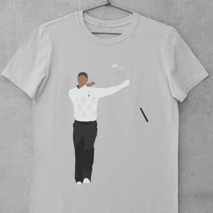 May include: Light gray t-shirt featuring a minimalist illustration of a golfer in a white sweater with a diamond pattern, black pants, and a blue cap. The golfer is holding a golf club. The shirt is hanging on a metal hanger.